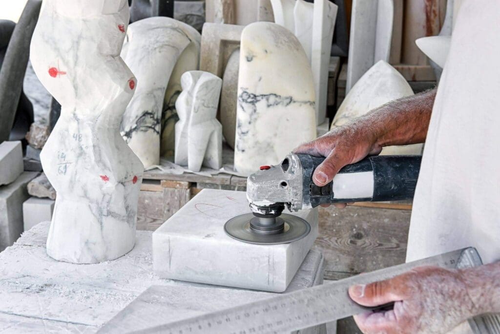 A person uses an angle grinder on a marble block in a workshop filled with abstract stone sculptures. A metal ruler is held in one hand, and marble dust covers the workspace and hands.