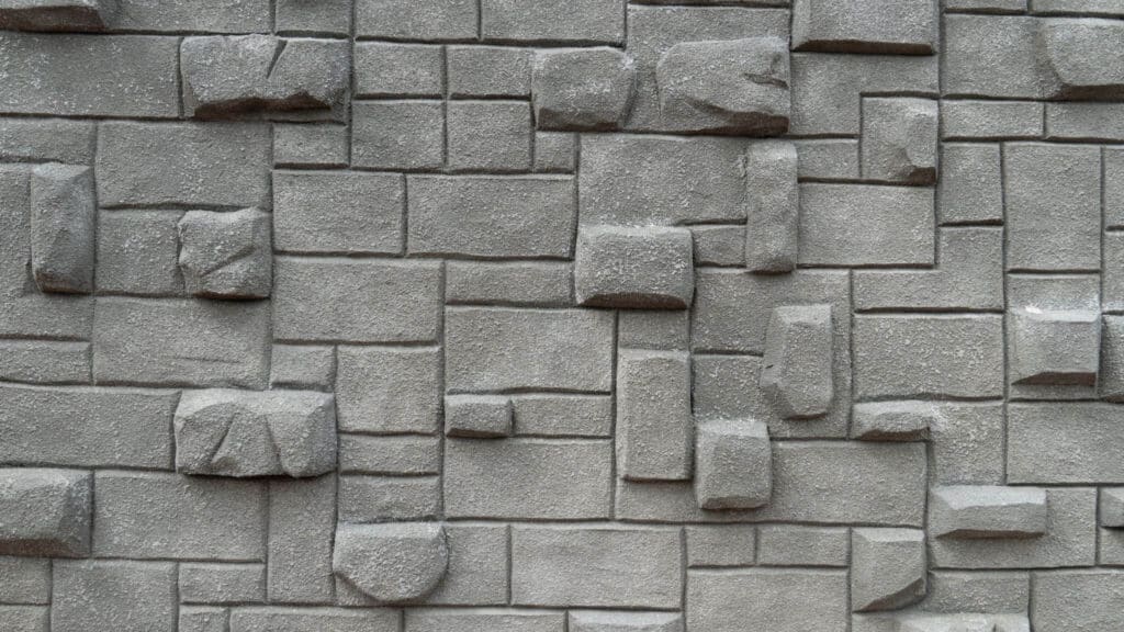 Gray textured concrete wall with a geometric pattern of rectangular and square blocks, some of which protrude, creating a three-dimensional effect.