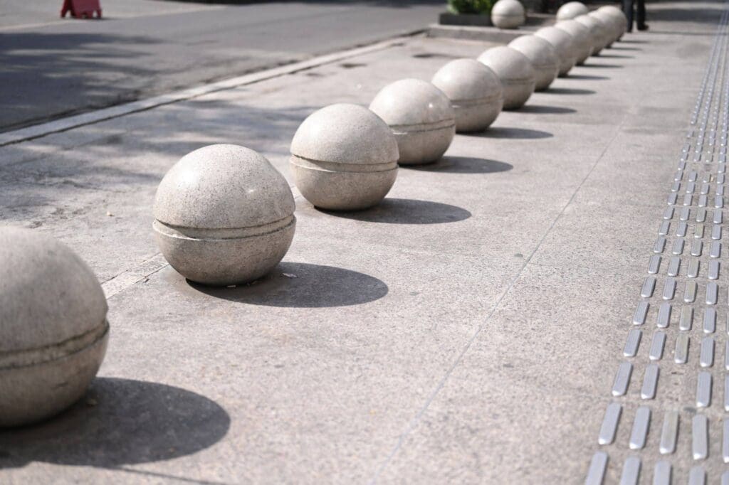 Concrete spheres are evenly spaced along a sidewalk next to a tactile paving strip. The spheres cast round shadows, and a road is visible in the background.