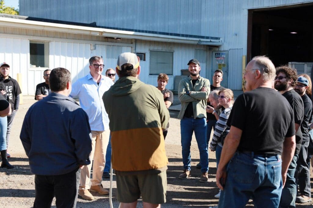 A group of people, including an architect and a stone fabricator, stand in a semi-circle outdoors near a building, listening to a man in a light blue shirt speak. Sunlight casts shadows as collaboration unfolds among the casually dressed gathering.