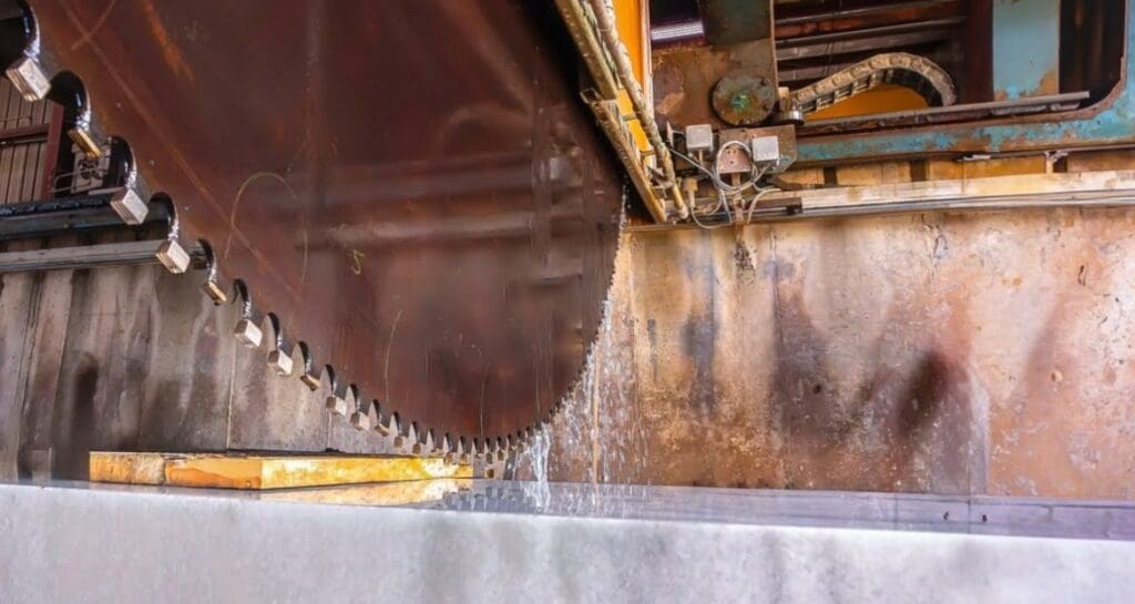 A large industrial saw blade cuts through a slab of stone or marble, with water spraying to cool the blade, as part of an architect and stone fabricator collaboration inside a factory setting.