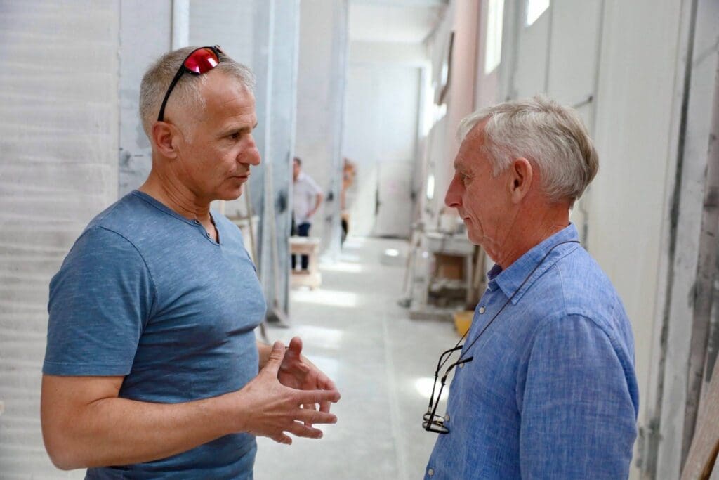 Two older men stand facing each other in conversation inside a bright, industrial-looking space. Their engaged gestures suggest an architect and stone fabricator collaboration, deep in discussion about their latest project.