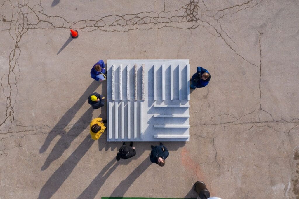Aerial view of six people standing around a rectangular concrete structure with steps, casting long shadows on cracked pavement. This scene hints at an architect and stone fabricator collaboration. An orange cone is visible in the top left corner.