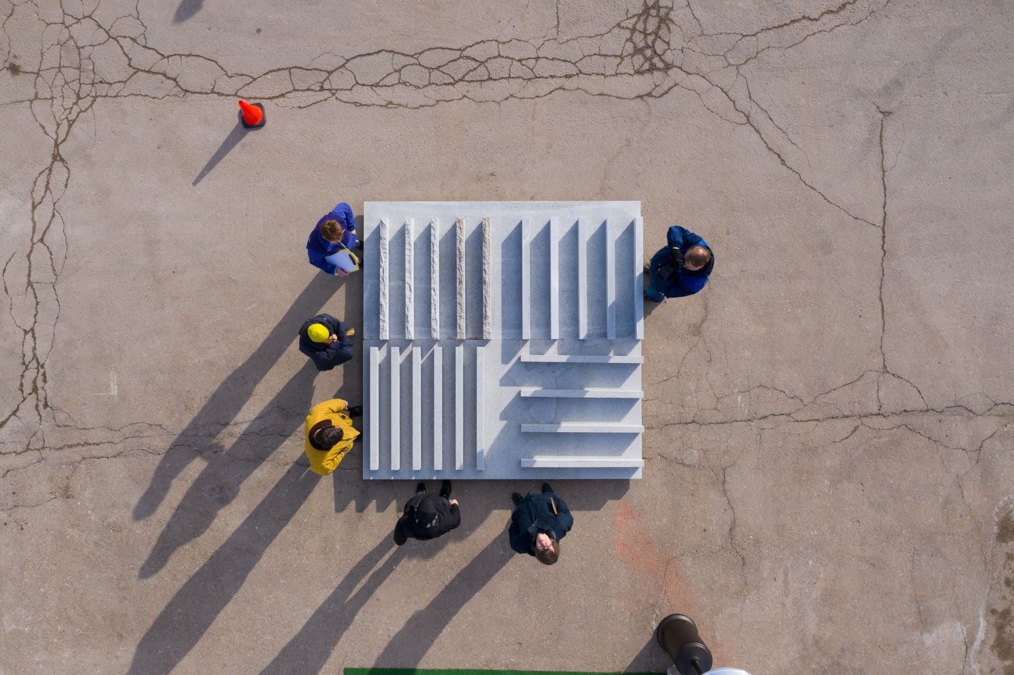 Aerial view of six people standing around a rectangular concrete structure with steps, casting long shadows on cracked pavement. This scene hints at an architect and stone fabricator collaboration. An orange cone is visible in the top left corner.