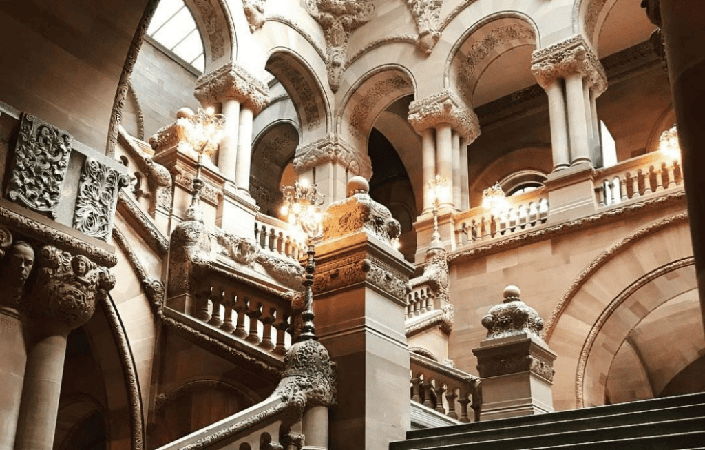 Ornate natural stone staircase with detailed carvings, grand arches, columns, and decorative balustrades inside a historic building, lit by ornate lamps—a stunning showcase for designers and architects alike.