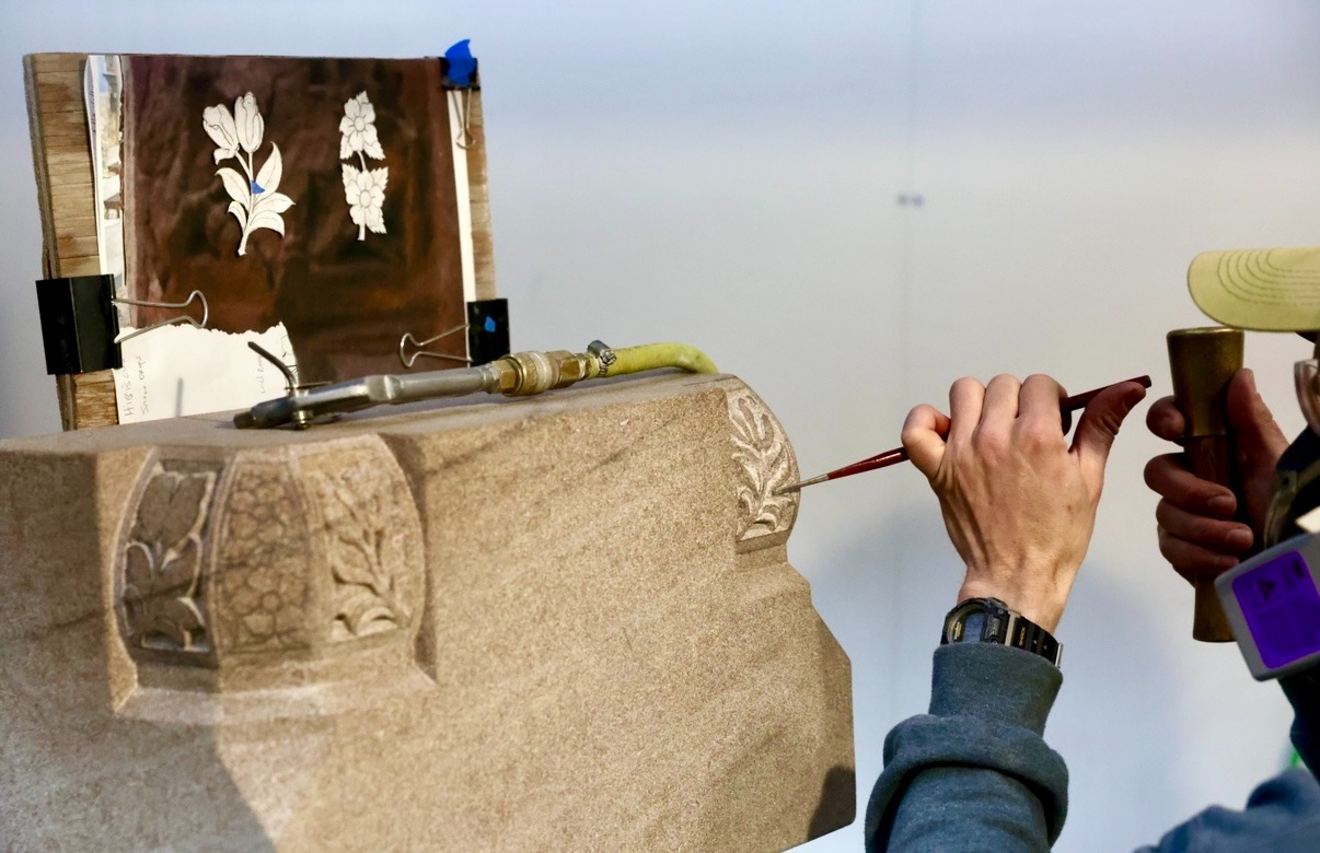 A person carves floral patterns into a stone block using a chisel and hammer, with a sheet of flower sketches attached to a board nearby for reference.