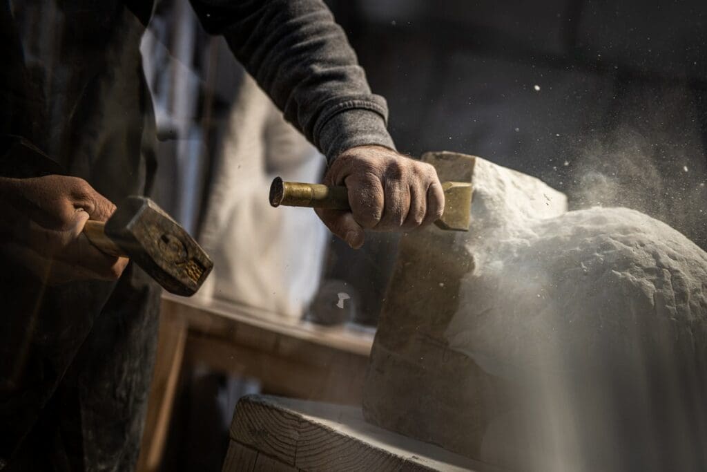 A person uses a hammer and chisel to carve stone, with dust rising from the impact. The focus is on their hands and the detailed motion of sculpting.