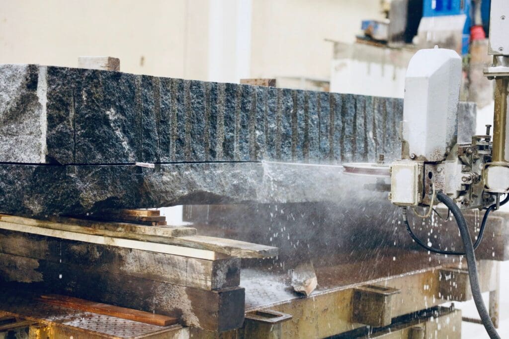 A stone-cutting machine sprays water as it slices through a large granite block, destined for Statement Stone Walls or other Architectural Surfaces, which rests on stacked wooden pallets in an industrial setting.