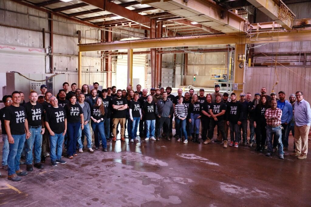 A large group of people pose for a photo inside an industrial warehouse, some wearing matching black T-shirts. The group stands on a wet concrete floor beneath metal beams and overhead cranes, surrounded by bold surfaces and striking architectural details.