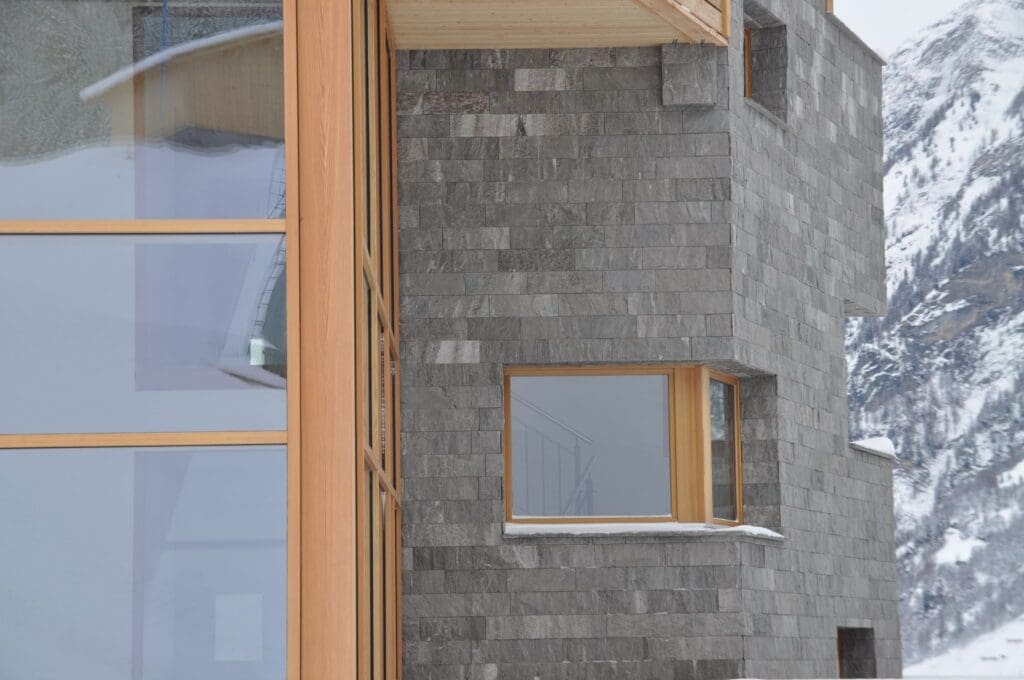 Modern building with gray stone walls and wooden-framed windows, set against a snowy mountain landscape. Part of the building reflects in the large glass windows on the left side.