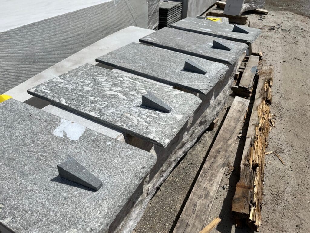 Six large rectangular stone slabs with smaller triangular stone pieces attached on top, arranged in a row on wooden planks outdoors, possibly at a construction or stone supply site.