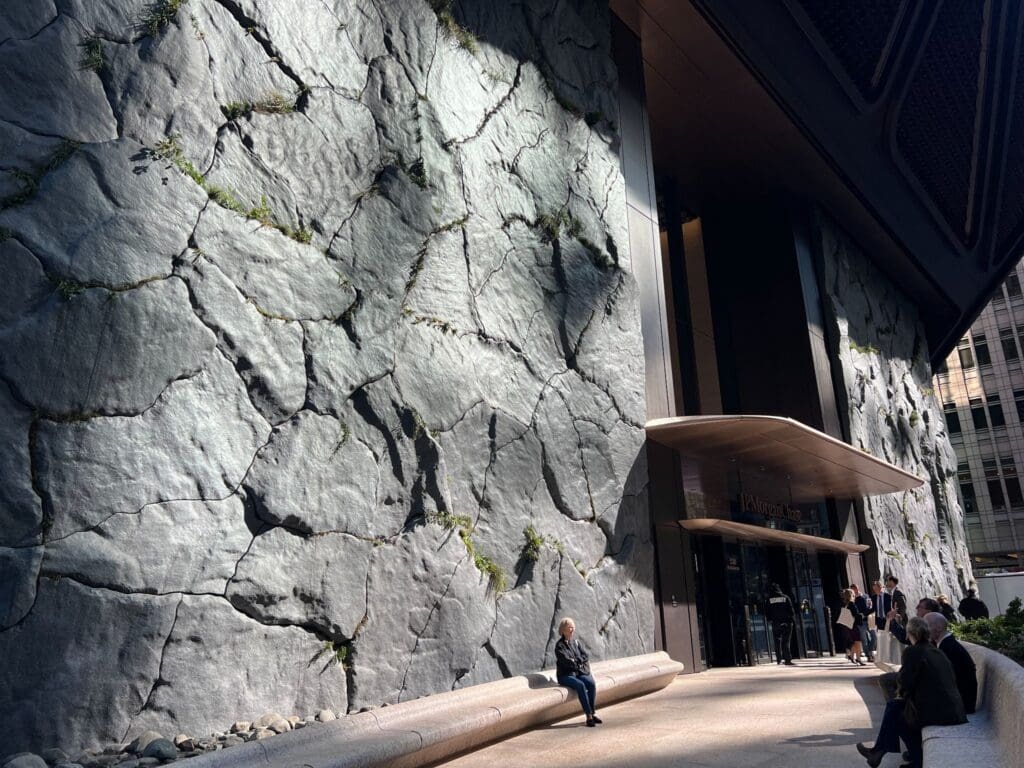 A person sits on a stone-textured wall outside a building entrance, with sunlight casting shadows. Several people stand and walk nearby.
