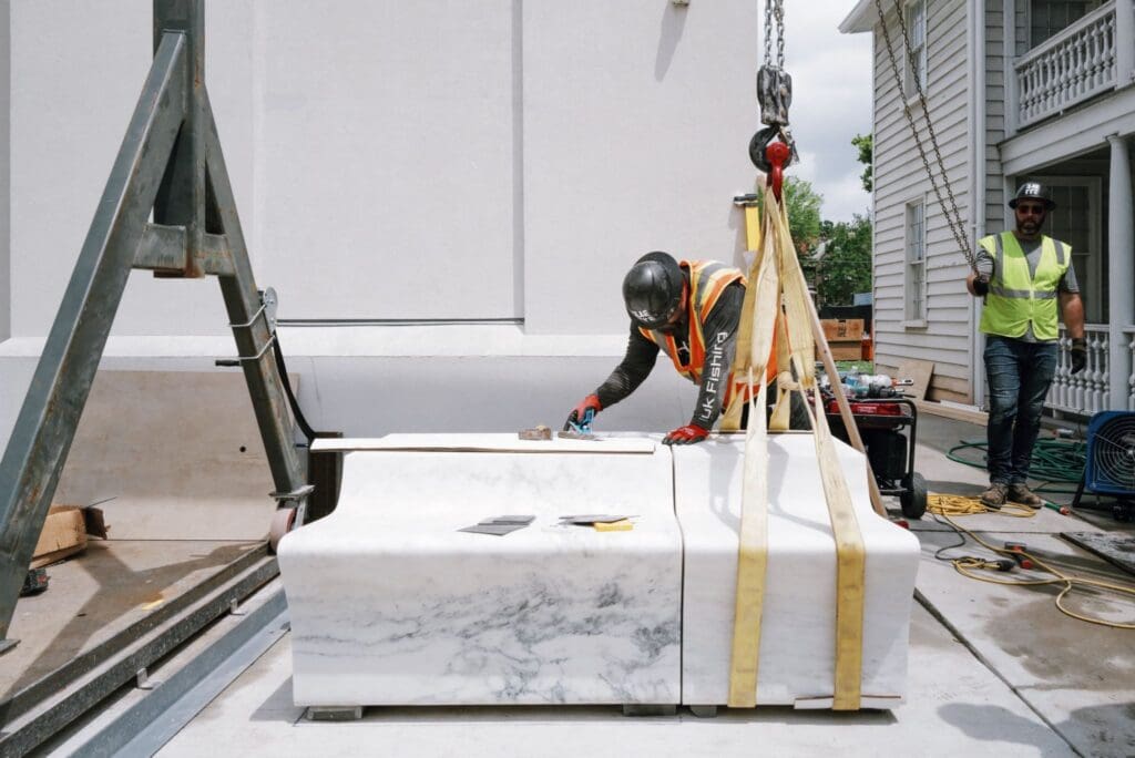 A construction worker in safety gear inspects a large marble countertop being lifted by a crane, while another worker stands nearby outside a white building.