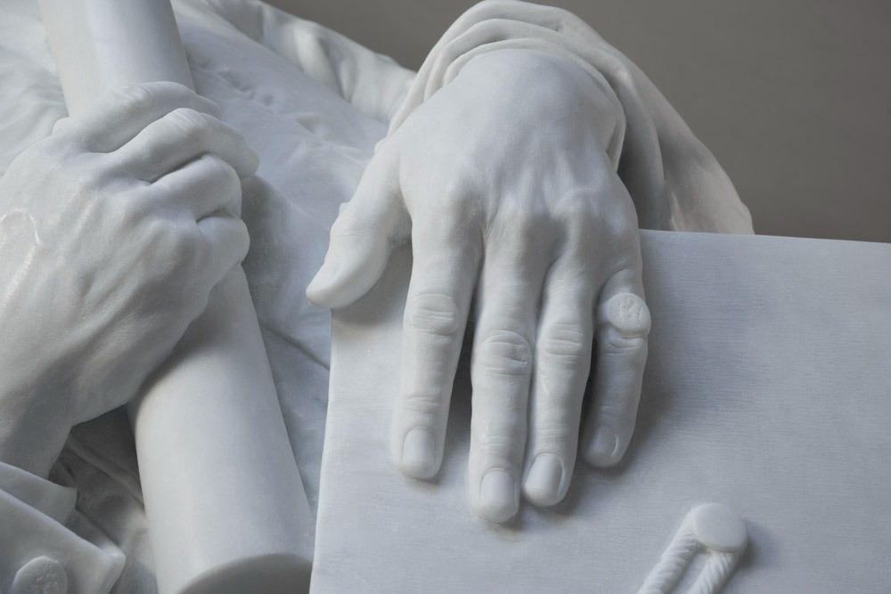 Close-up of a marble statue showing one hand holding a diploma and the other resting on a graduation cap with a tassel.