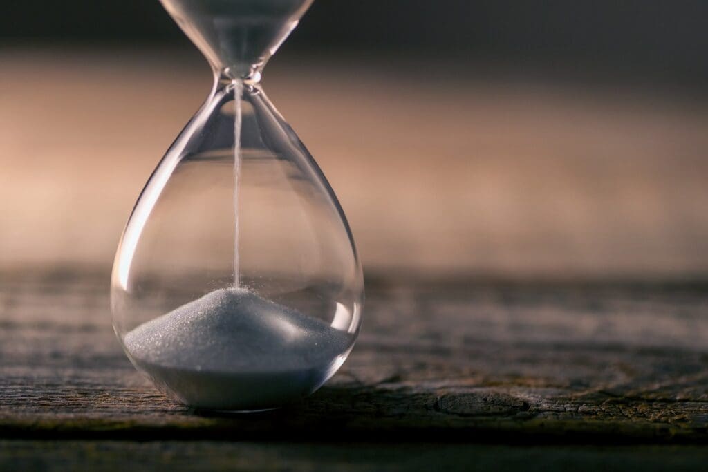 A close-up of an hourglass with white sand flowing down, placed on a wooden surface with a blurred background.