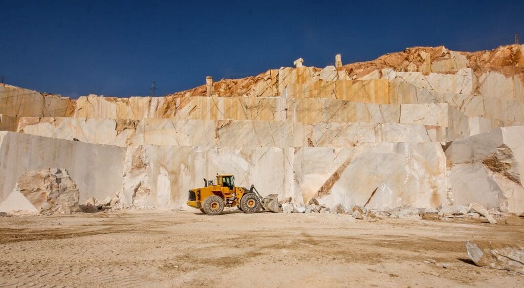 A yellow bulldozer is parked on sandy ground in front of large, terraced marble quarry walls under a clear blue sky.