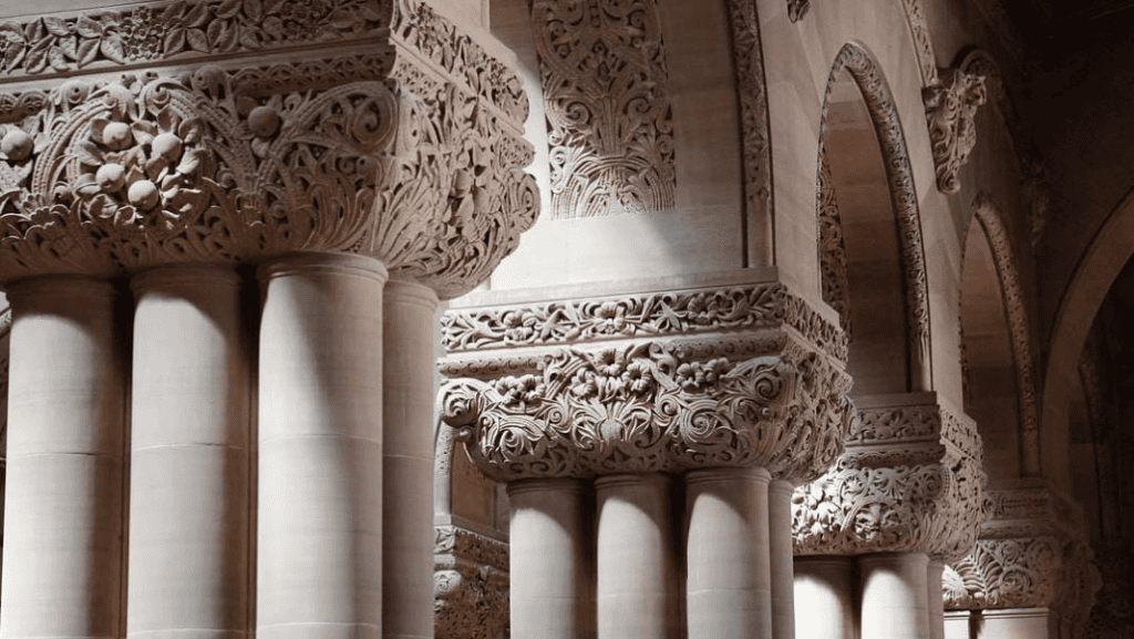 Ornate natural stone columns and arches with intricate floral carvings are illuminated by soft light inside a historic building, showcasing the craftsmanship cherished by builders and architects alike.