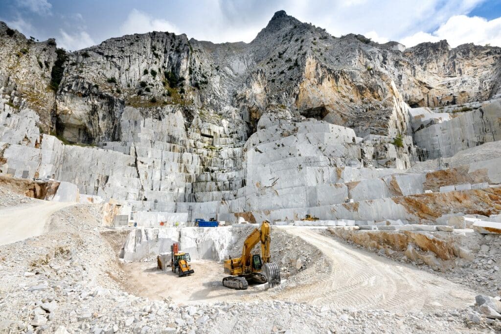 A marble quarry with steep white stone walls, heavy machinery, and excavators at the base. Rocky mountains rise in the background under a partly cloudy sky.