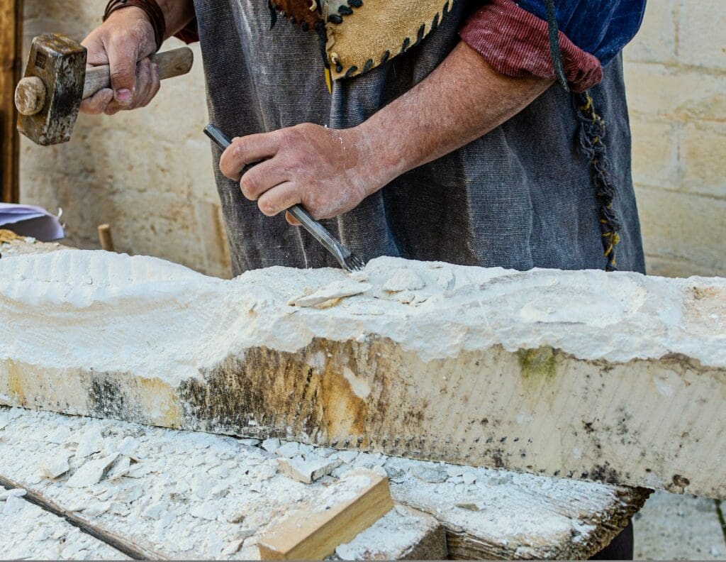 A person uses a hammer and chisel to carve into a long slab of white stone, wearing a leather apron and working on a table covered in stone dust and small wood pieces.
