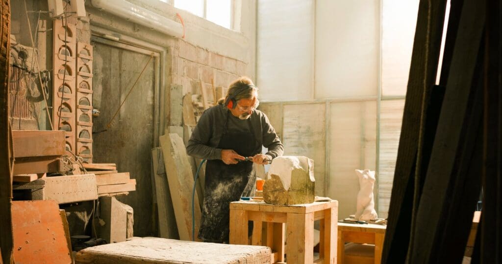 An artist in a sunlit workshop carves a stone sculpture on a wooden table, surrounded by various tools, stone blocks, and unfinished pieces.