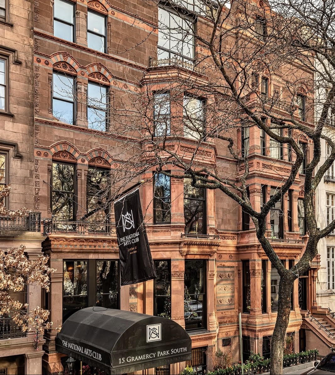 A brownstone building with large windows and a black awning labeled 15 GRAMERCY PARK SOUTH stands proudly, its entrance distinguished by a flag bearing The National Arts Club logo. Leafless trees frame the scene, enhancing the distinguished atmosphere of this iconic cultural venue.