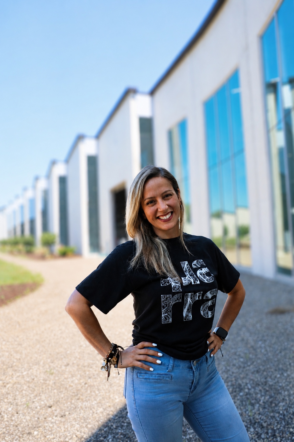Smiling woman with long blonde hair stands outside a modern glass building, wearing a black graphic t-shirt and blue jeans, with hands on hips on a sunny day—celebrating August Work Anniversaries at Quarra Stone Company.