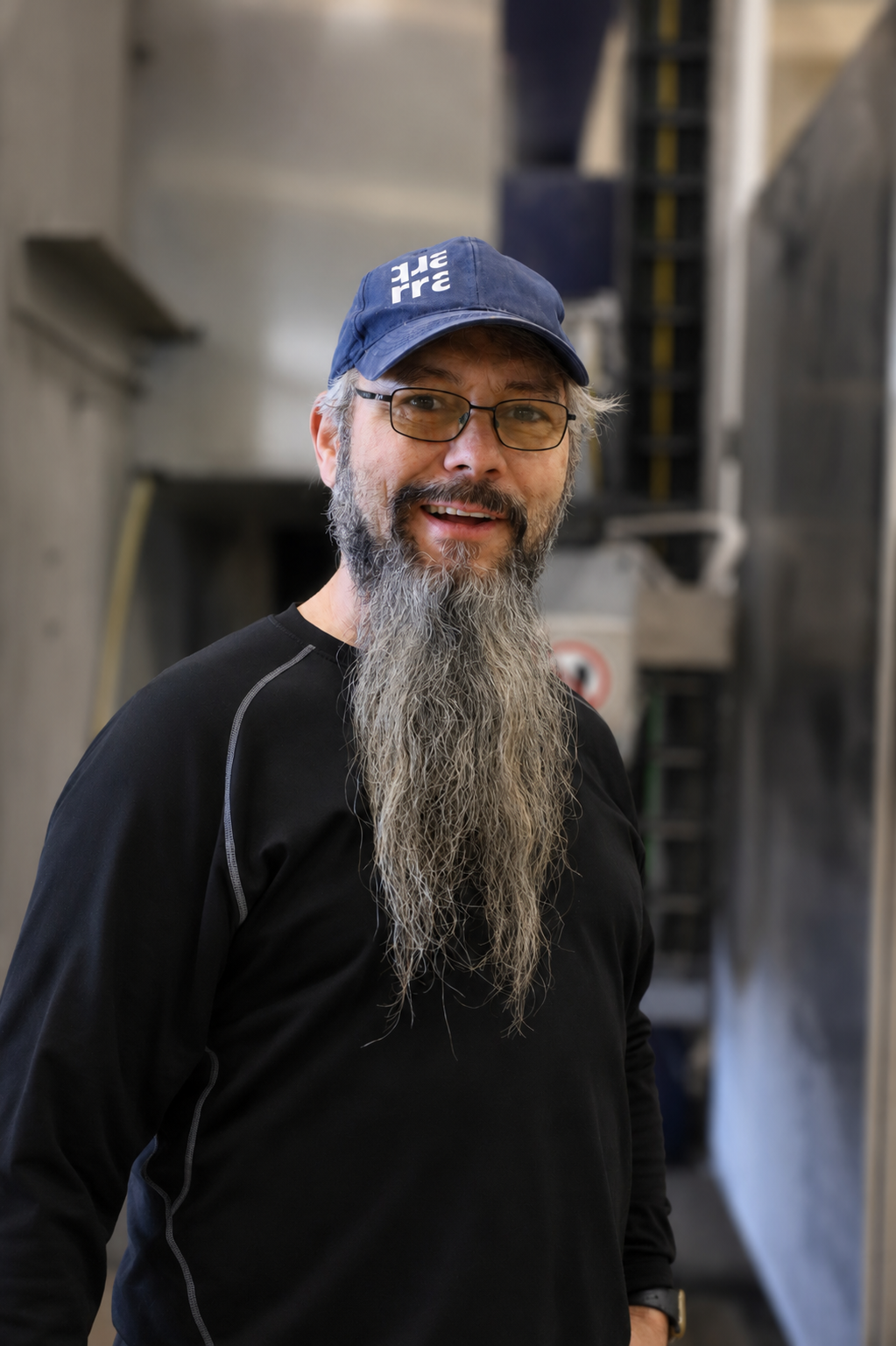 A man with glasses and a long gray beard smiles, wearing a blue cap and black shirt, standing indoors in an industrial setting at Quarra Stone Company during the August Work Anniversaries celebration.