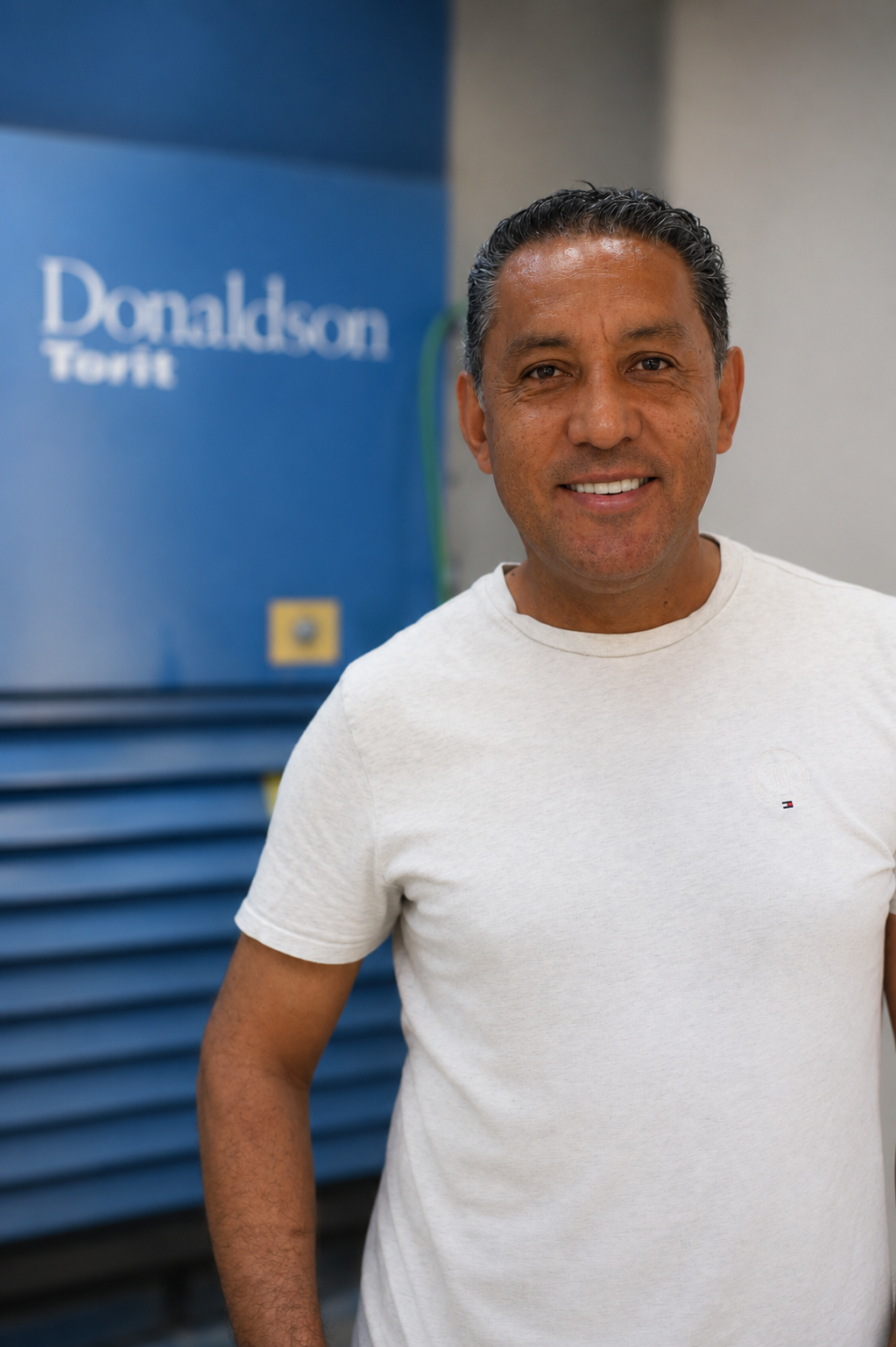 A smiling, middle-aged man with dark hair wears a white t-shirt. He is standing in front of a large, blue industrial machine with Donaldson Torit labeled on the side, proudly Celebrating August Work Anniversaries at Quarra Stone Company.