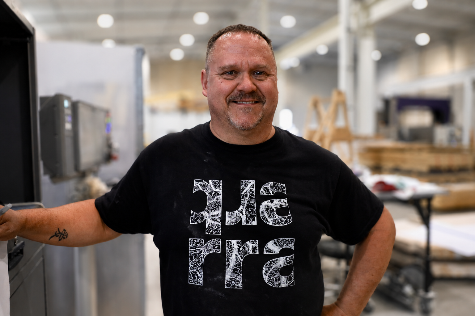 A man with short hair and a goatee smiles while standing in a bright, industrial workspace at Quarra Stone Company. He wears a black T-shirt with the word Quarra in large, white letters on the front, celebrating August work anniversaries among his dedicated team.